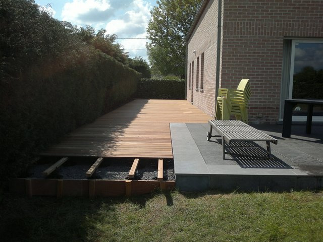 Drewniany taras w trakcie budowy, widoczne deski, podkonstrukcja i fragment wykończonej części z leżakiem na tle ceglanego domu i żywopłotu.