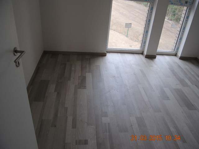 Wnętrze pokoju z nową, jasną podłogą panelową i dwoma oknami PCV w białych ramach. Widoczny fragment drzwi z metalową klamką po lewej stronie.