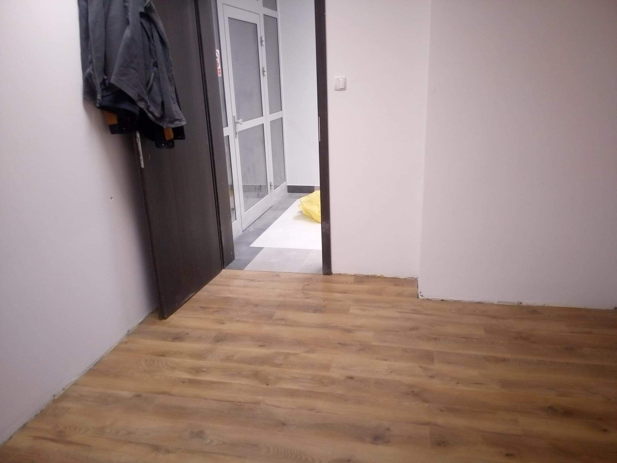 Widok na świeżo położoną podłogę panelową w korytarzu, z widoczną częścią drzwi wejściowych z kurtką zawieszoną na klamce i białymi drzwiami na końcu korytarza.