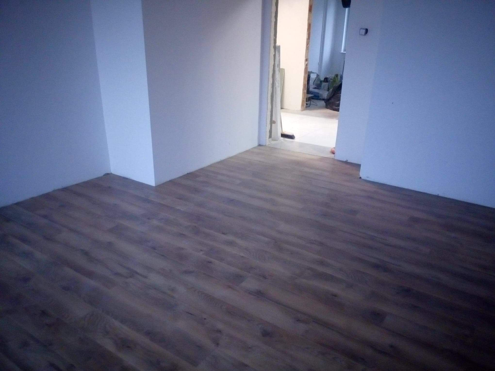 Nowo położona, ciemna podłoga panelowa w pustym, odmalowanym na biało pokoju, widoczny otwór drzwiowy do kolejnego pomieszczenia.