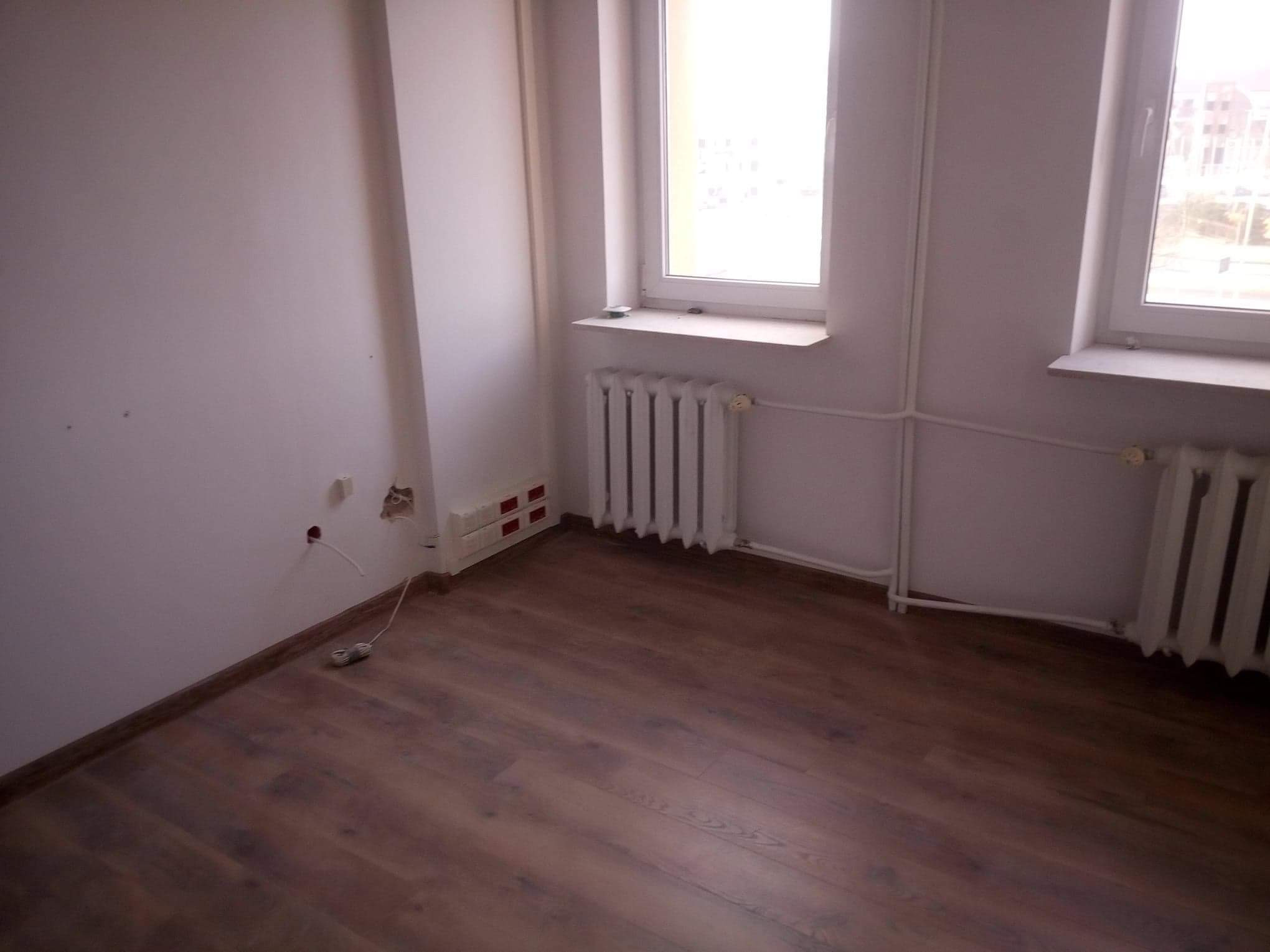 Puste pomieszczenie z dwoma oknami, grzejnikiem i panelami podłogowymi w odcieniach brązu, widoczne niedokończone prace elektryczne przy ścianie.