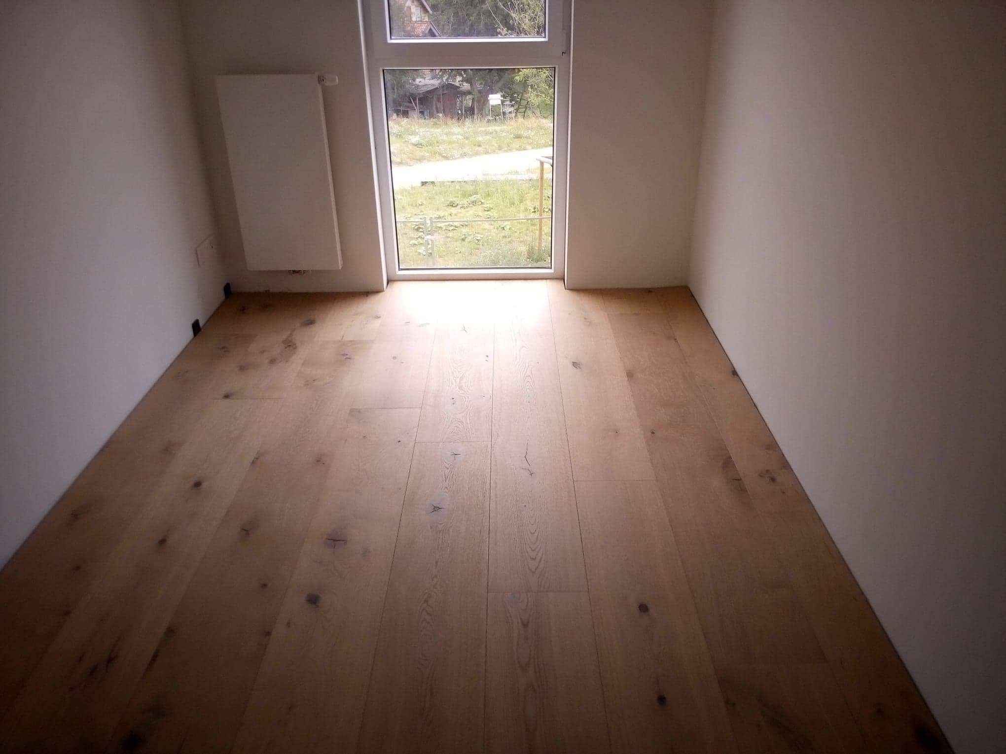 Pusta przestrzeń z nową drewnianą podłogą, świeżo pomalowanymi ścianami i oknem z widokiem na podwórko.