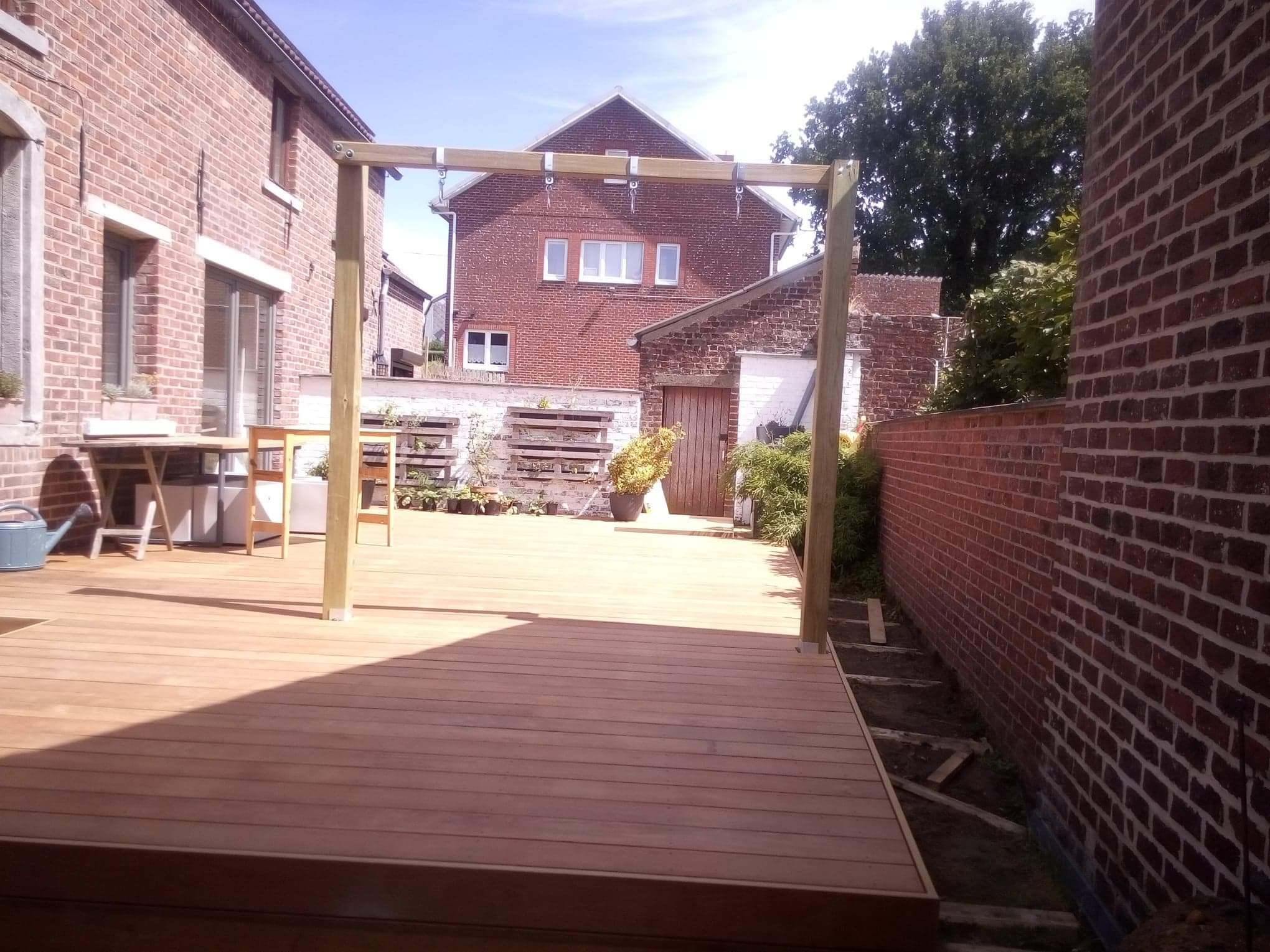 Nowo wybudowany drewniany taras z pergolą w ogrodzie, otoczony ceglanymi budynkami i zielenią.