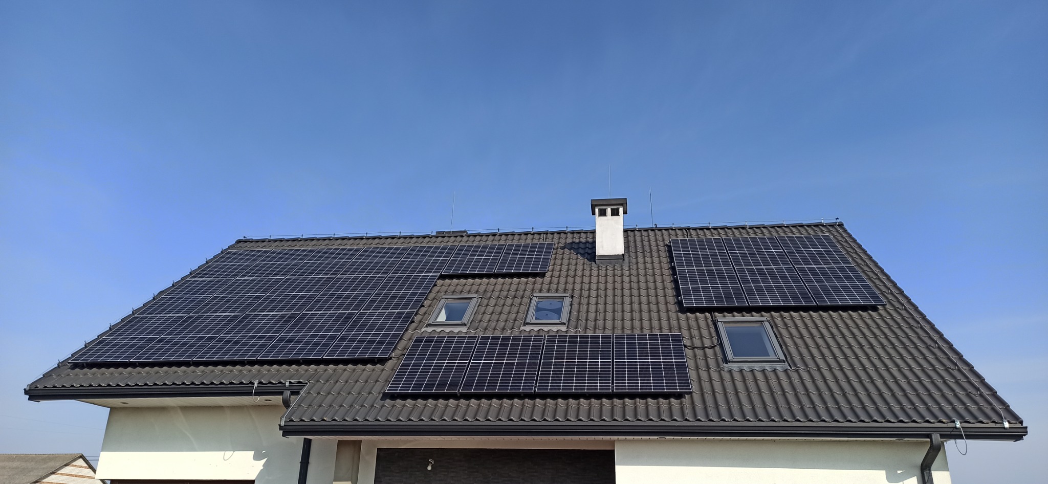 Panele słoneczne na dachu domu z dachówką ceramiczną, widoczne okna dachowe i komin, błękitne niebo w tle.