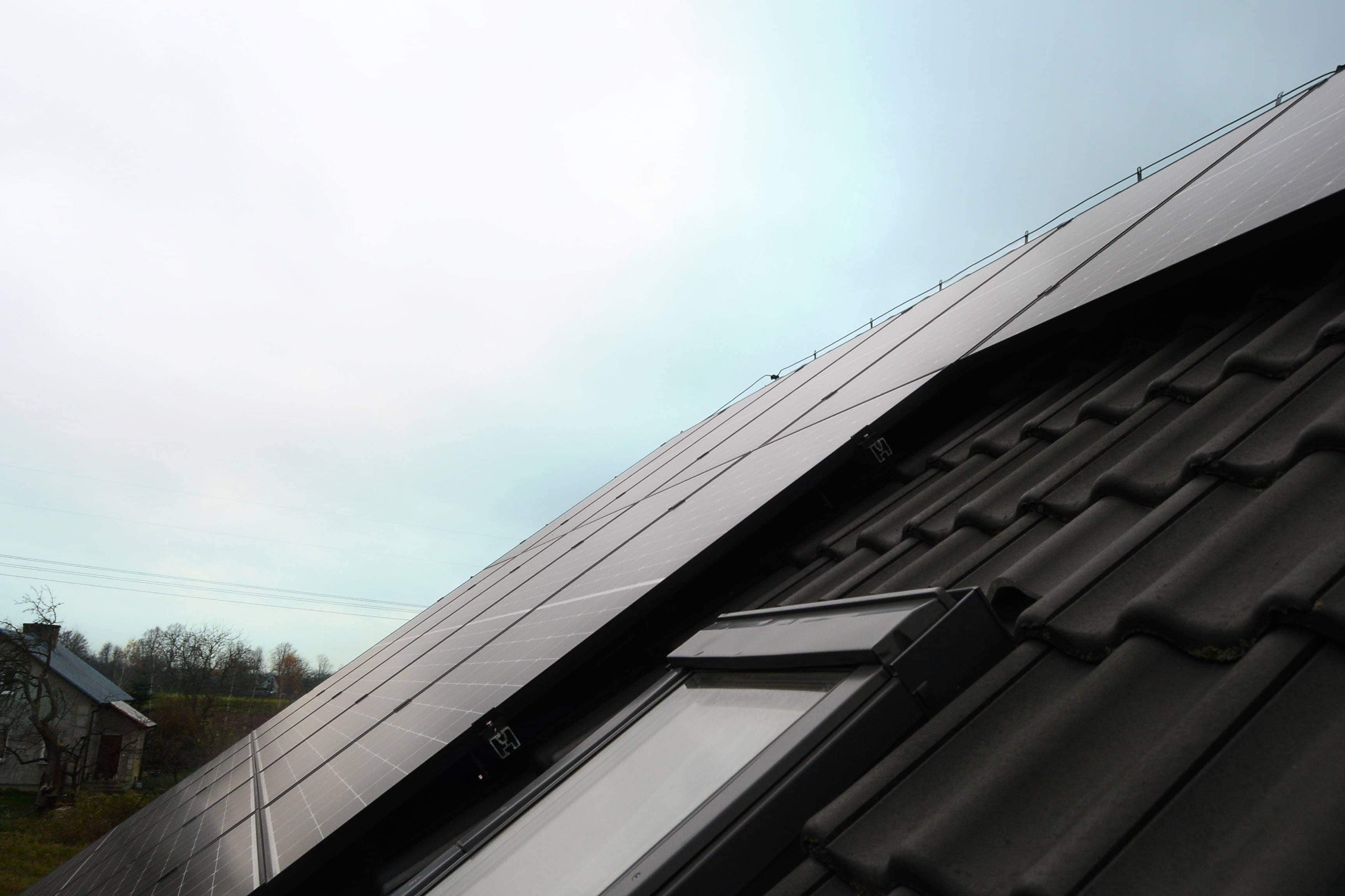 Czarny dach z dachówkami i oknem dachowym, na którym zamontowane są panele fotowoltaiczne w pochmurny dzień, widoczny fragment budynku w tle.