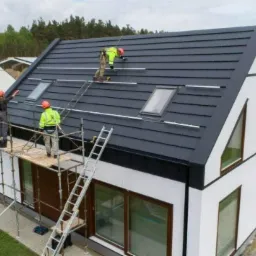 Montaż konstrukcji pod panele fotowoltaiczne na dachu nowoczesnego domu z oknami dachowymi; ekipa w kaskach i odblaskowych kamizelkach pracuje na rusztowaniu i drabinie.