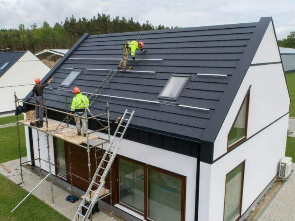 Montaż konstrukcji pod panele fotowoltaiczne na dachu nowoczesnego domu z oknami dachowymi; ekipa w kaskach i odblaskowych kamizelkach pracuje na rusztowaniu i drabinie.
