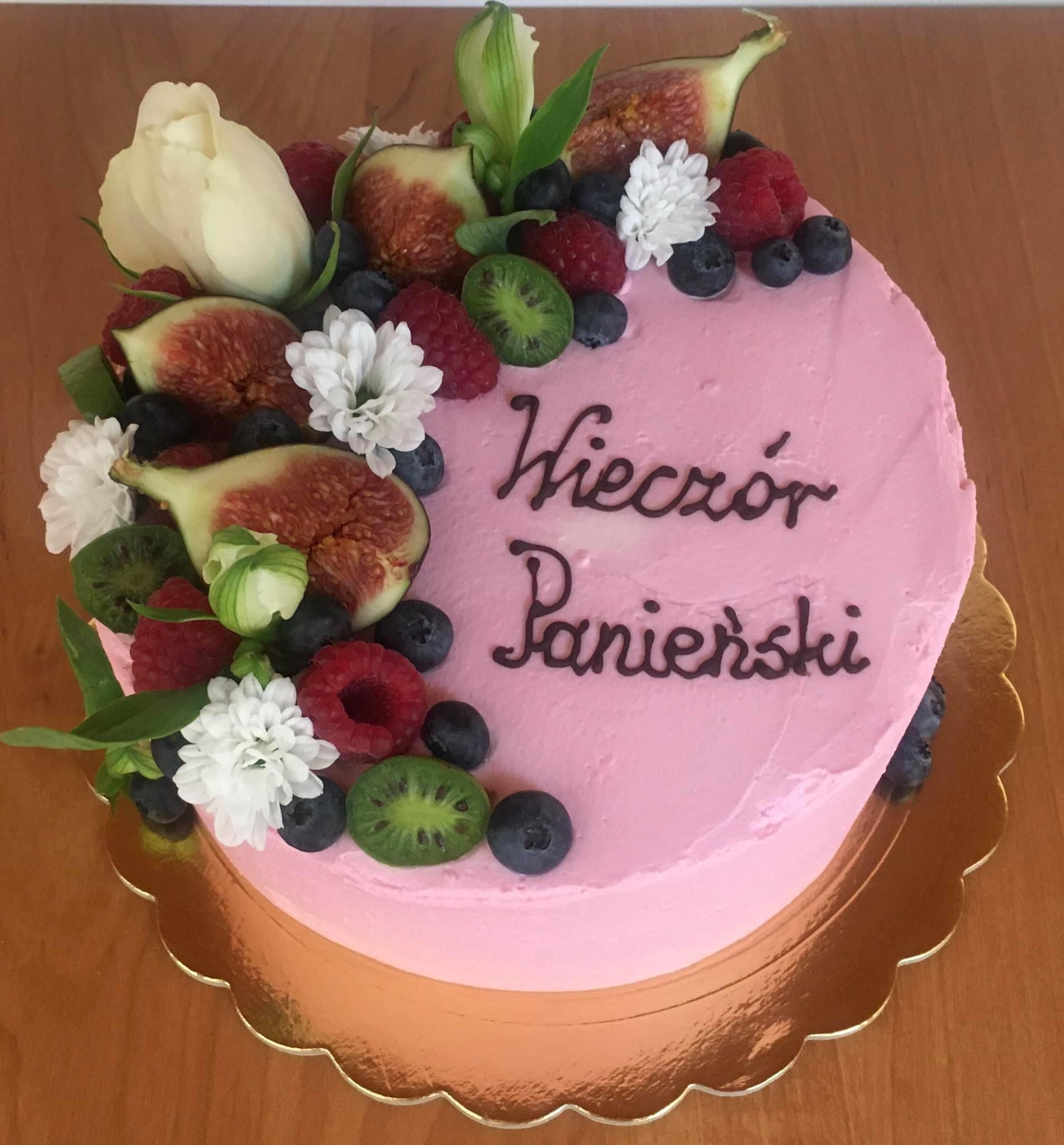 Różowy tort z napisem 'Wieczór Panieński', udekorowany figami, malinami, borówkami, kiwi, białymi kwiatami i białą różą, umieszczony na złotej, ozdobnej podkładce.