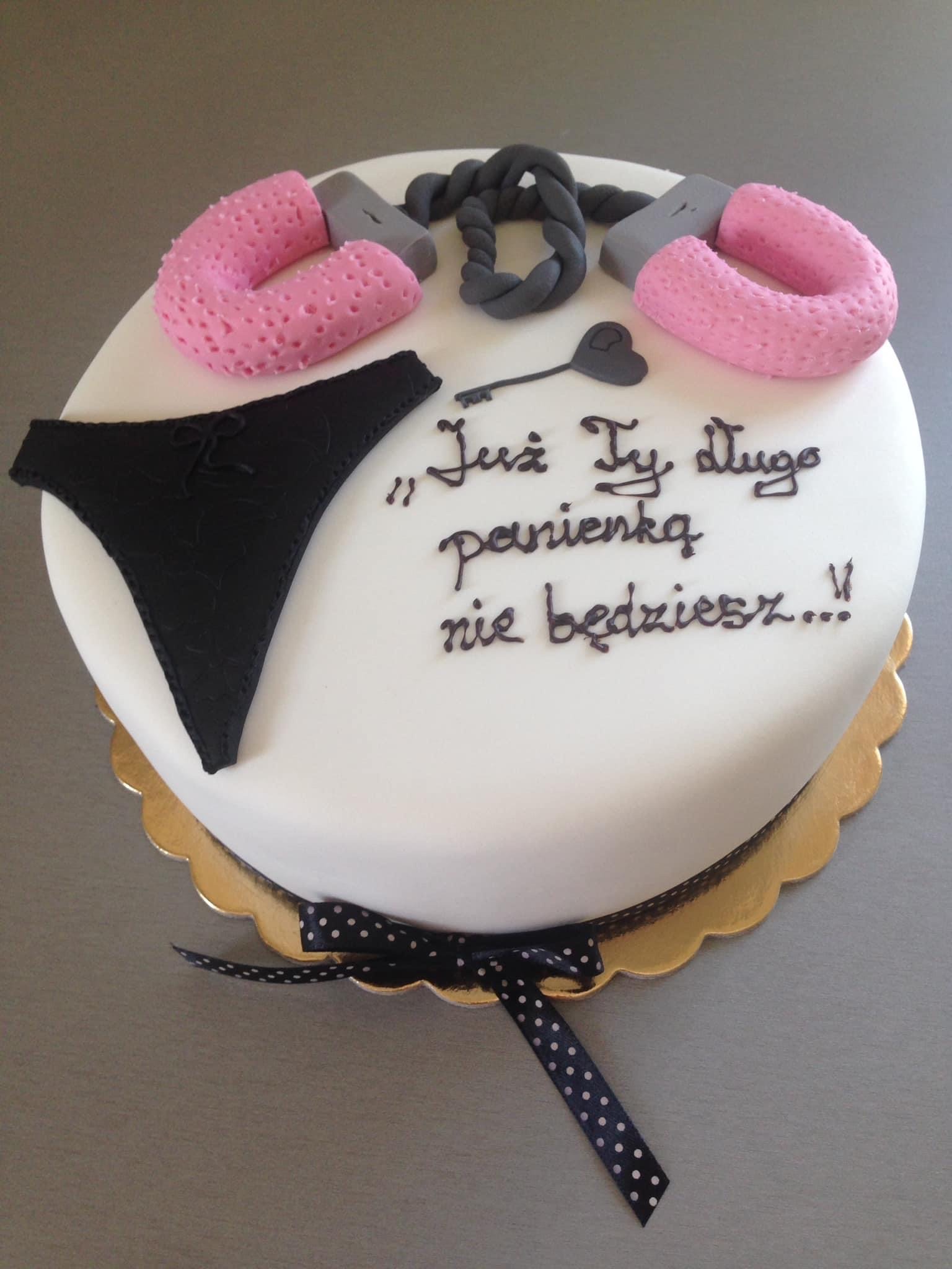 Okrągły tort z białym lukrem, ozdobiony czarną bielizną, różowymi kajdankami, szarym sznurem i napisem 'Już Ty długo panienką nie będziesz...!'. Pod tortem złota podkładka i czarna wstążka w białe...