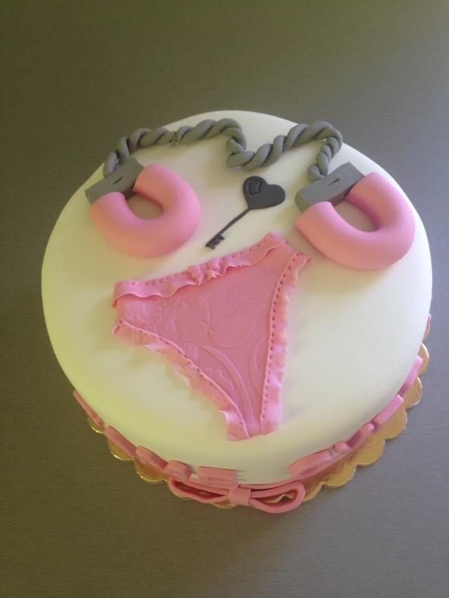 Okrągły tort z białym lukrem, ozdobiony modelowanymi elementami z masy cukrowej: różowe majtki z falbankami, szare kajdanki połączone sznurkiem oraz czarny klucz w kształcie serca.