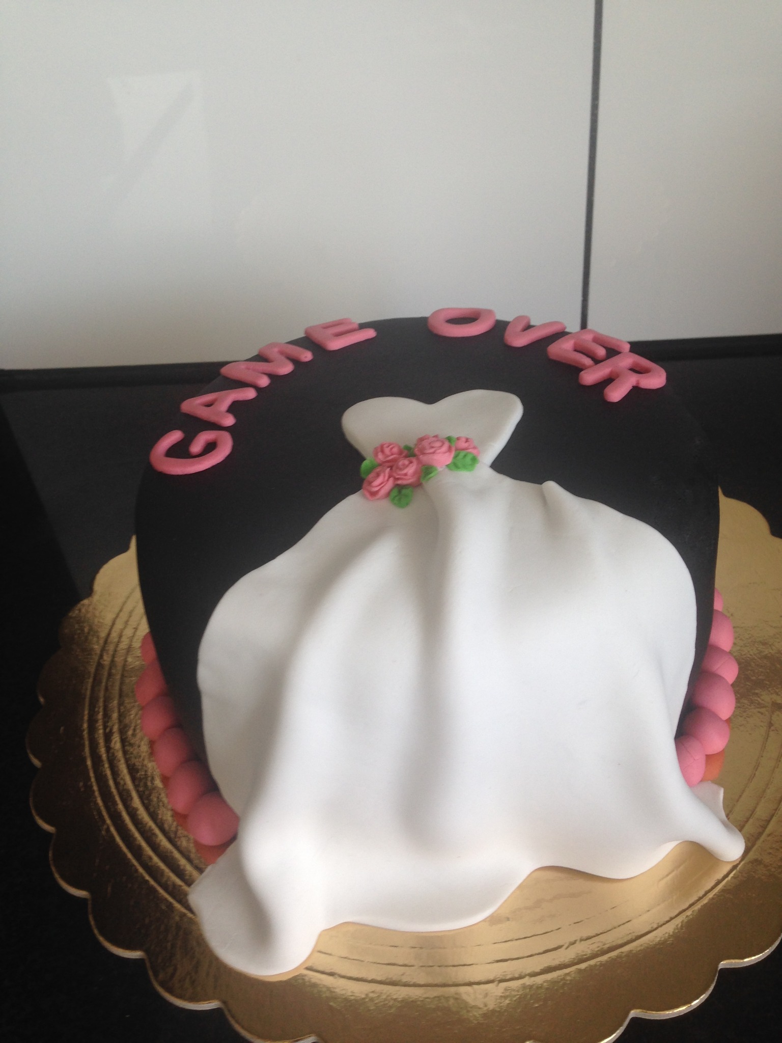 Czarny tort z napisem 'Game Over' i dekoracją w kształcie białej sukni ślubnej z różowymi różyczkami, umieszczony na złotej podkładce.