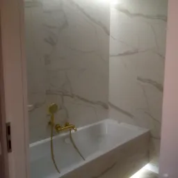 Nowoczesna łazienka z wanną obudowaną marmurowymi płytkami, złotą armaturą i podświetleniem LED pod wanną i nad nią.