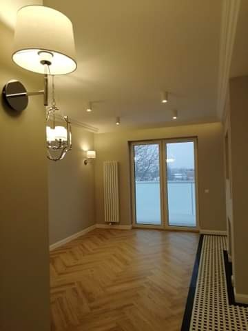 Jasne, nowocześnie wykończone wnętrze z parkietem w jodełkę, widoczne drzwi balkonowe i ozdobne oświetlenie ścienne.