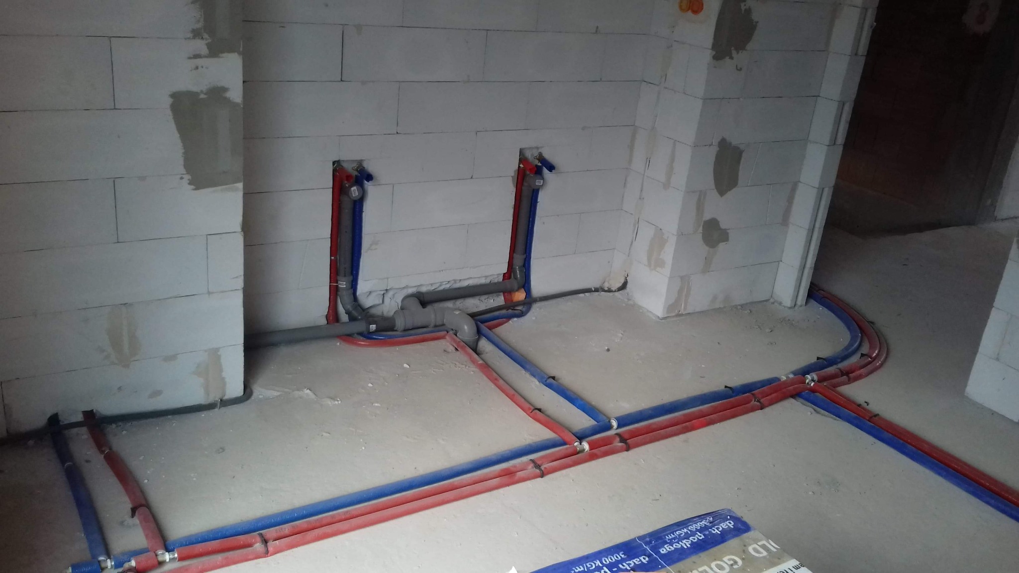 Wnętrze w trakcie prac hydraulicznych: widoczne czerwone i niebieskie rury prowadzone po podłodze i w ścianach z betonu komórkowego, szare rury kanalizacyjne, instalacja w surowym stanie.