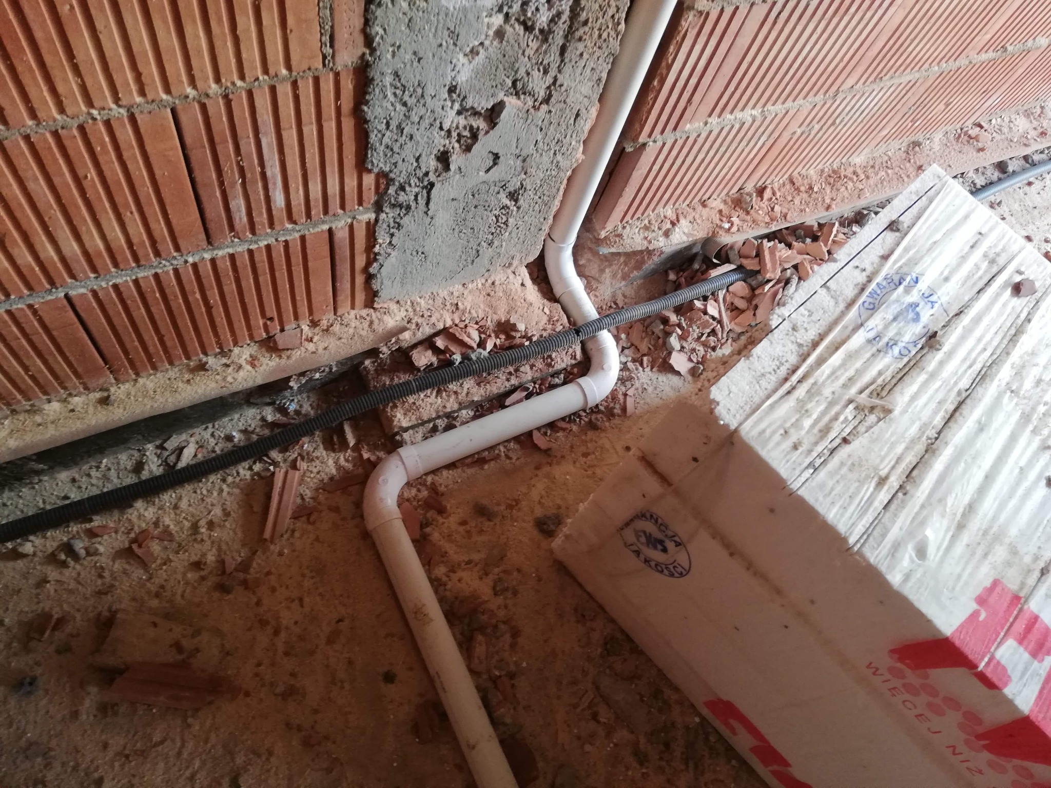 Białe rury instalacji wodnej ułożone wzdłuż murowanej ściany z cegły dziurawki w trakcie prac wykończeniowych, widoczne także czarne rury karbowane i kartonowe opakowanie