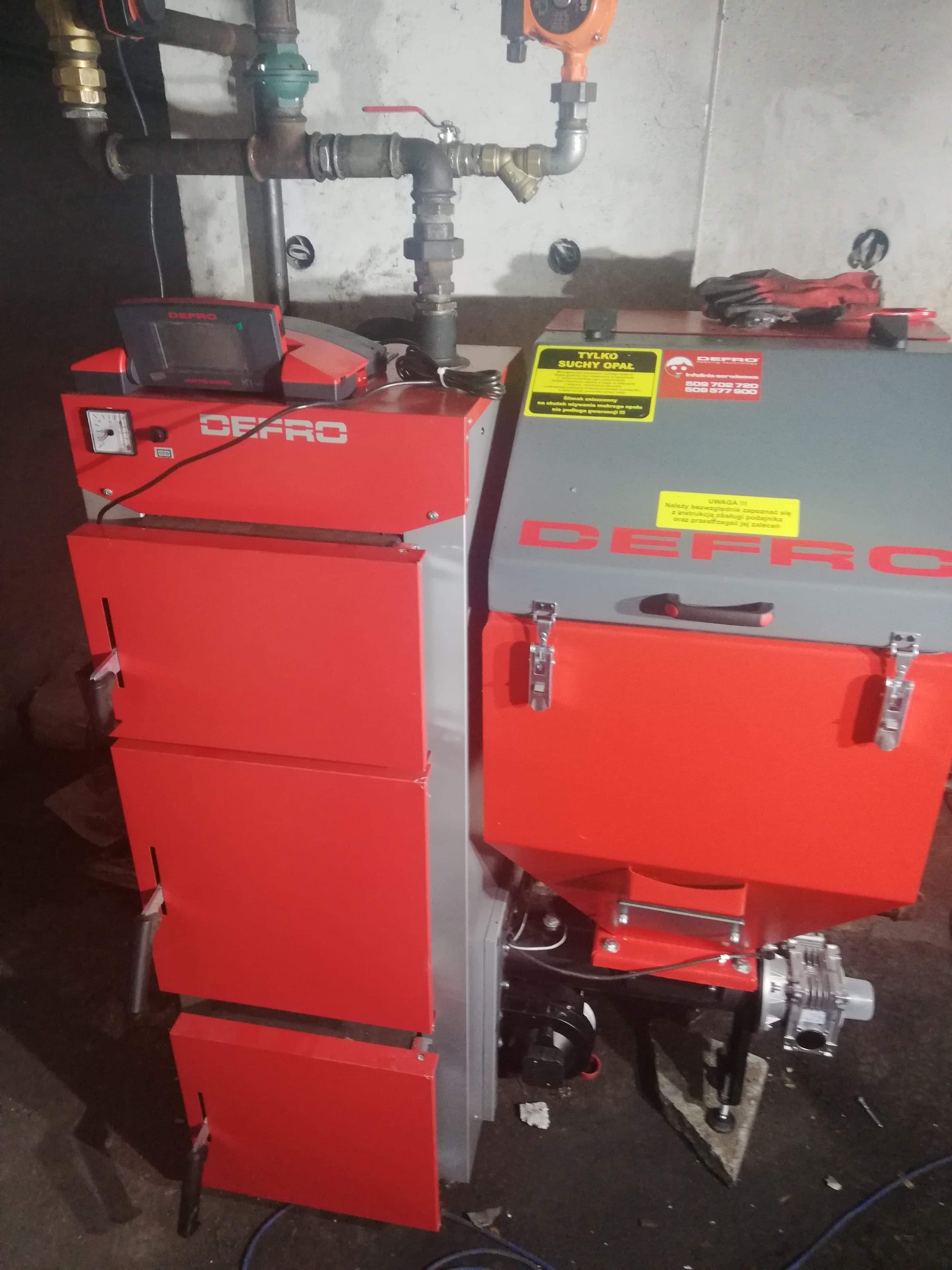 Nowy, czerwono-szary piec CO marki Defro z podajnikiem, stojący w kotłowni, widoczne rury instalacji centralnego ogrzewania i tabliczki informacyjne na obudowie.