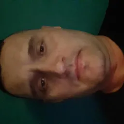 Portret mężczyzny z krótkimi włosami, skierowany w lewo, na zielonym tle. Widoczne rysy twarzy, skupiony wzrok. Obrócone ujęcie.