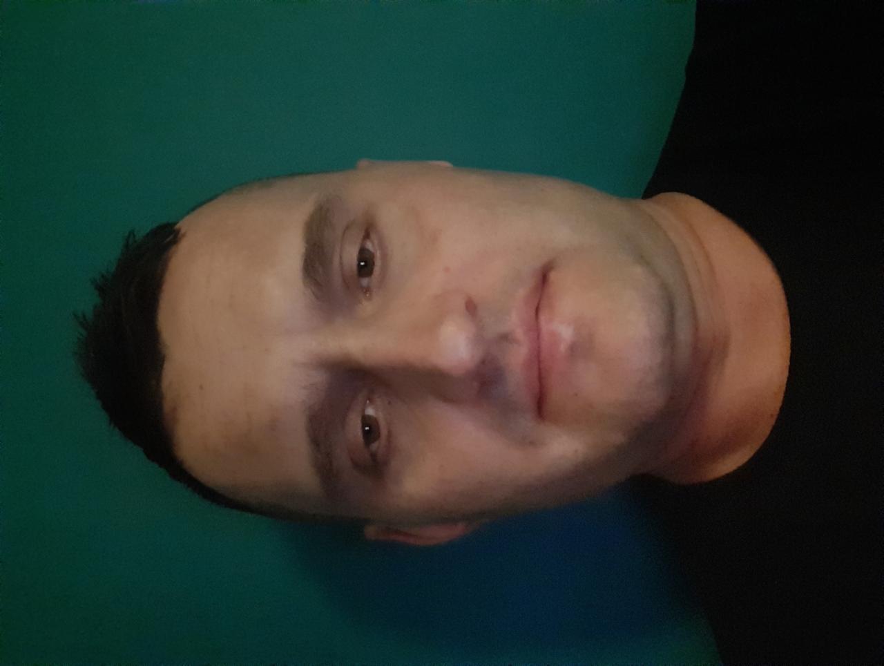 Portret mężczyzny z krótkimi włosami, ujęcie z bliska, z delikatnym uśmiechem, na tle jednolitej, zielonej ściany. Obrócony kadr.