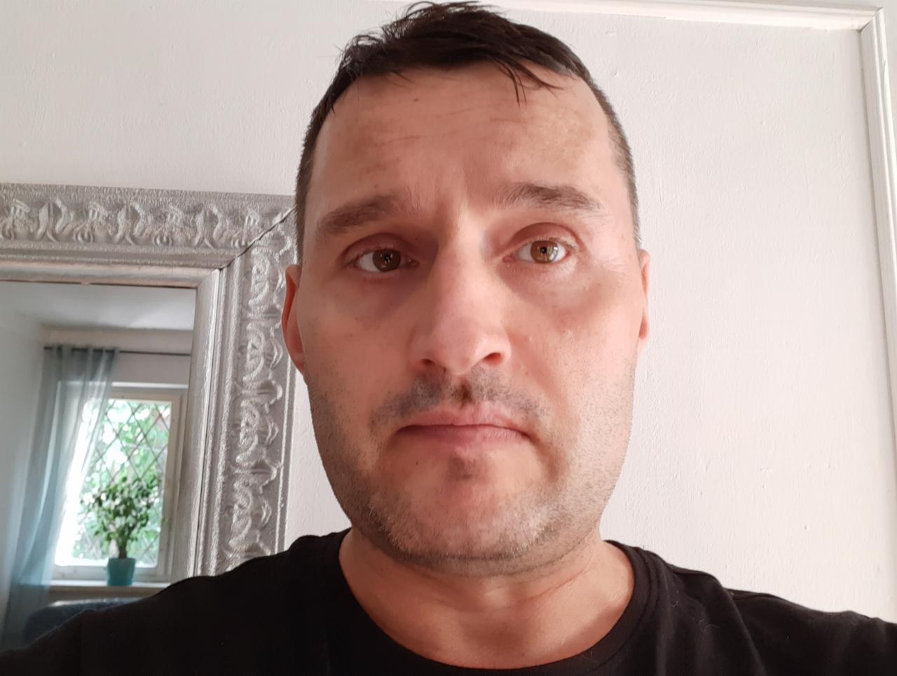 Portret mężczyzny w czarnej koszulce, z lustrem w ozdobnej ramie i rośliną w tle.