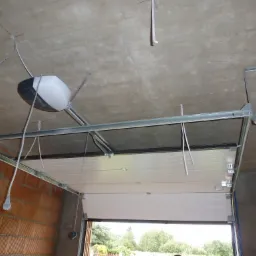 Widok od dołu na montaż napędu do bramy garażowej segmentowej z widocznym mechanizmem, szynami, kablem zasilającym i częściowo otwartą bramą, surowy betonowy strop i ściany z cegły.