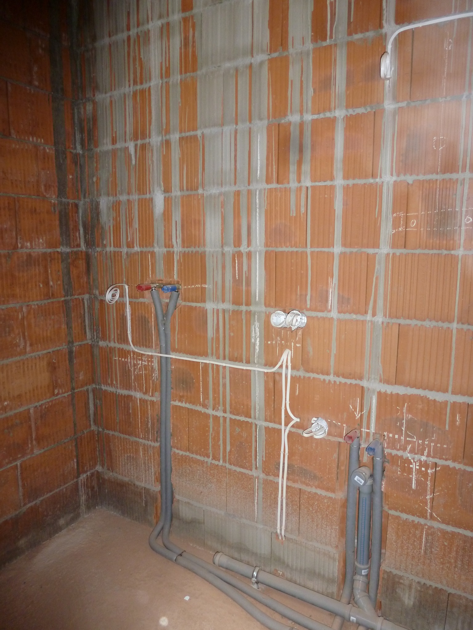 Surowy mur z czerwonej cegły z widocznymi instalacjami elektrycznymi i hydraulicznymi w trakcie montażu, w tym rurami z zaworami oznaczonymi kolorami niebieskim i czerwonym.