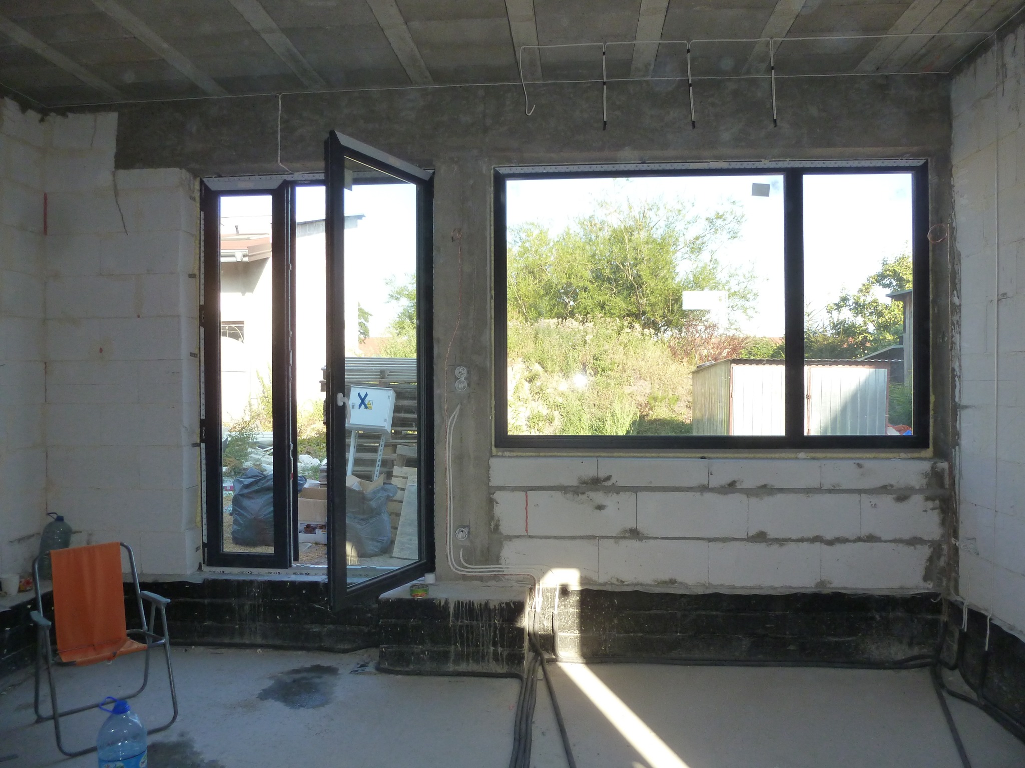 Surowy stan wnętrza z widocznymi przewodami elektrycznymi, nowymi oknami z czarnymi ramami i otwartymi drzwiami balkonowymi, ściany z białych bloczków, betonowy sufit, widoczne okablowanie...