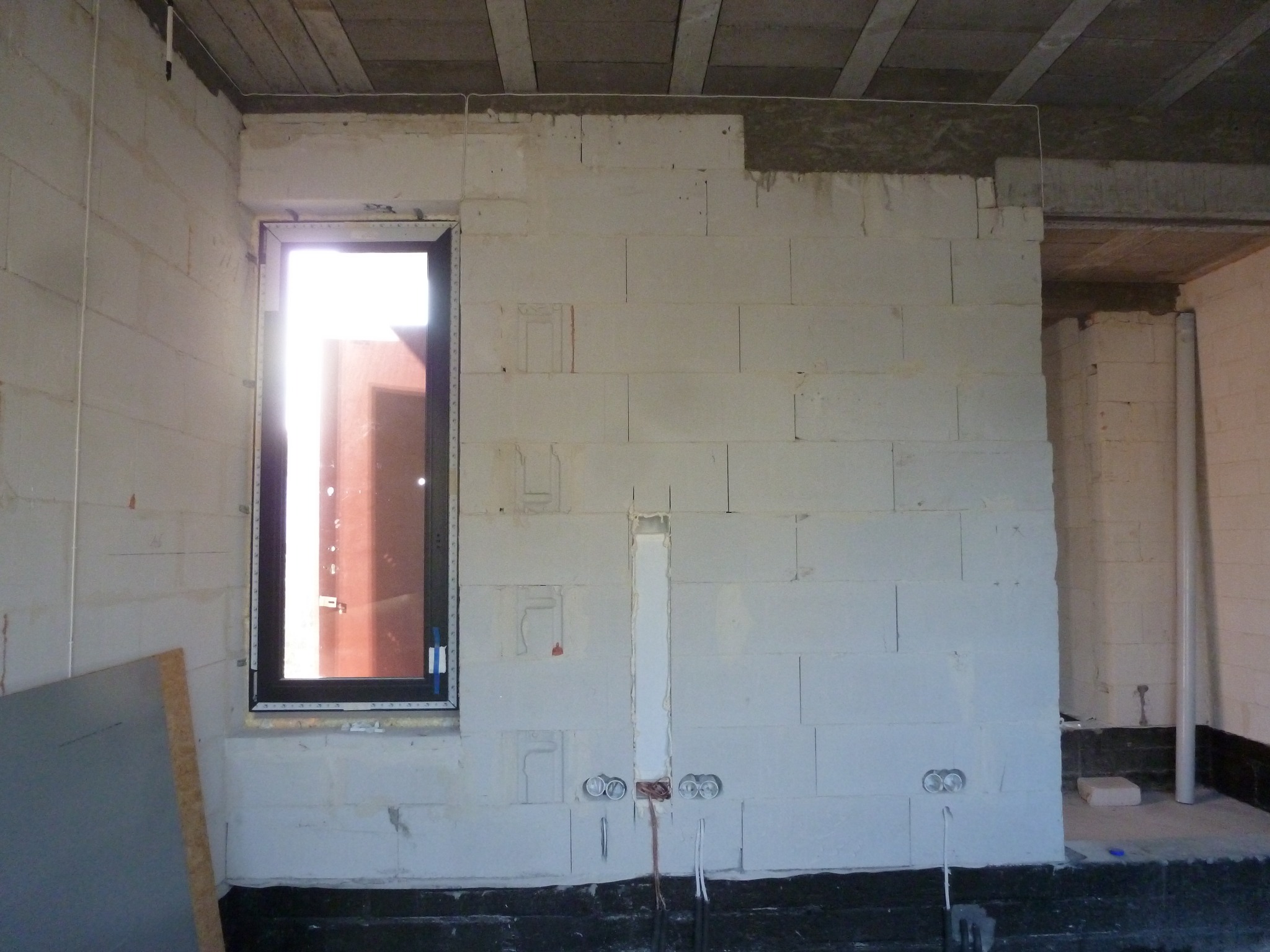 Surowy mur z betonu komórkowego z widocznymi puszkami elektrycznymi, przewodami i przygotowanymi bruzdami pod instalacje. Okno z czarną ramą w tle, widoczny fragment drewnianego stropu.