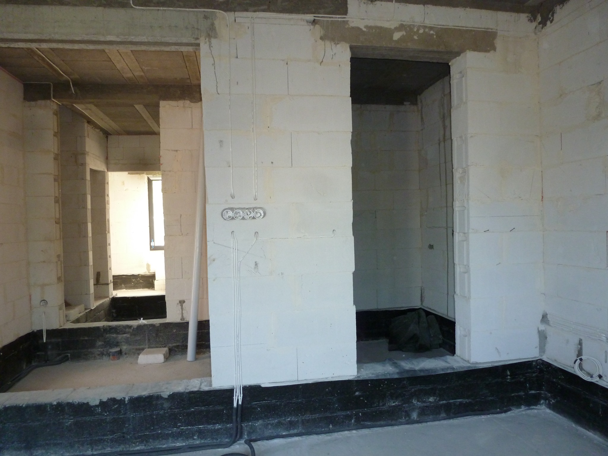 Surowy stan wnętrza budynku z białymi ścianami z betonu komórkowego, widoczne rury instalacji elektrycznej w ścianie i czarna izolacja przeciwwilgociowa przy podłodze.