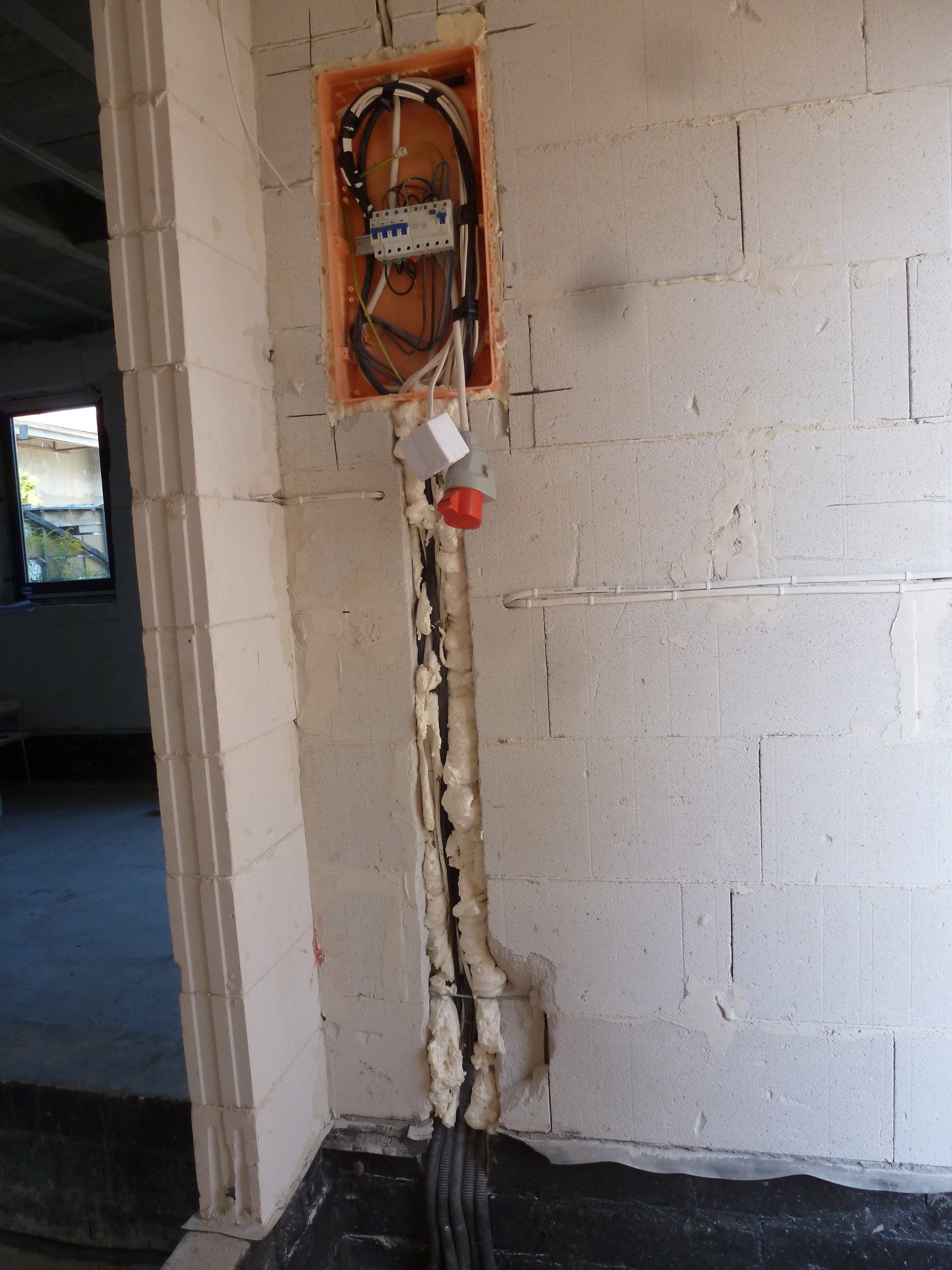 Wewnętrzna instalacja elektryczna w trakcie montażu: widoczna puszka z zabezpieczeniami, przewody w ścianie zabezpieczone pianką montażową, gniazdo siłowe i peszle z kablami przy podłodze, ściana...