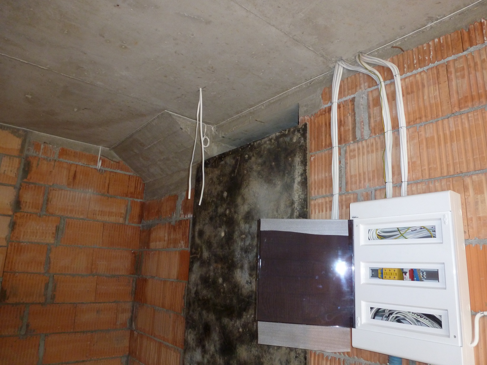 Otwarta, biała skrzynka elektryczna z widocznymi przewodami w surowym wnętrzu z ceglanymi ścianami i betonowym sufitem.