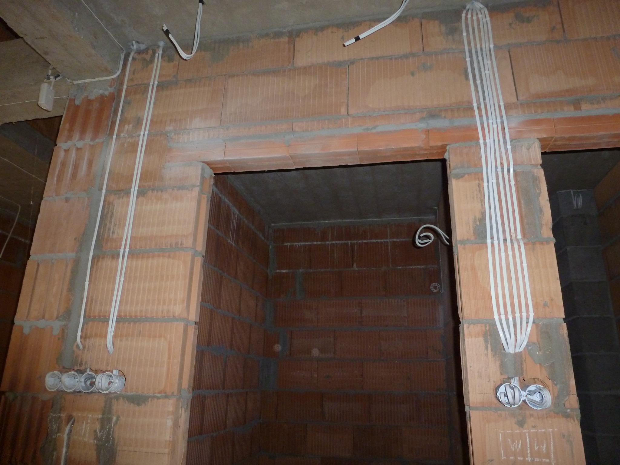 Instalacja elektryczna w surowym stanie: kable prowadzone po ceglanej ścianie, puszki pod gniazdka częściowo zamurowane, widoczne przewody w peszlach.