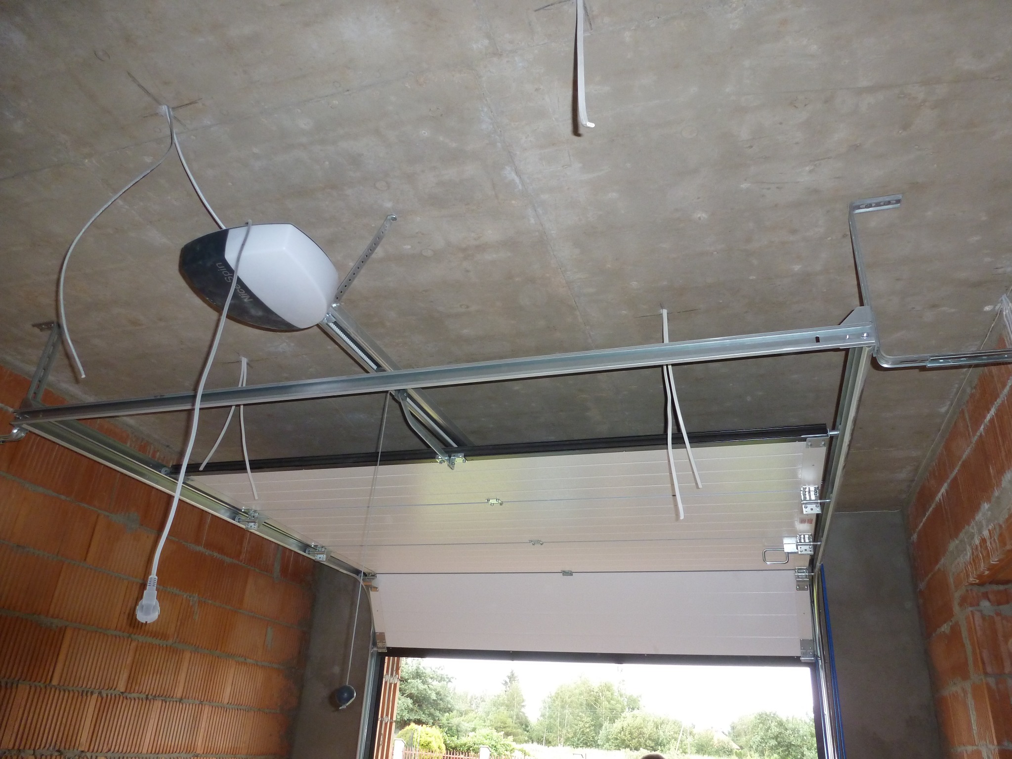 Widok od dołu na montaż napędu do bramy garażowej segmentowej z widocznym mechanizmem, szynami, kablem zasilającym i częściowo otwartą bramą, surowy betonowy strop i ściany z cegły.