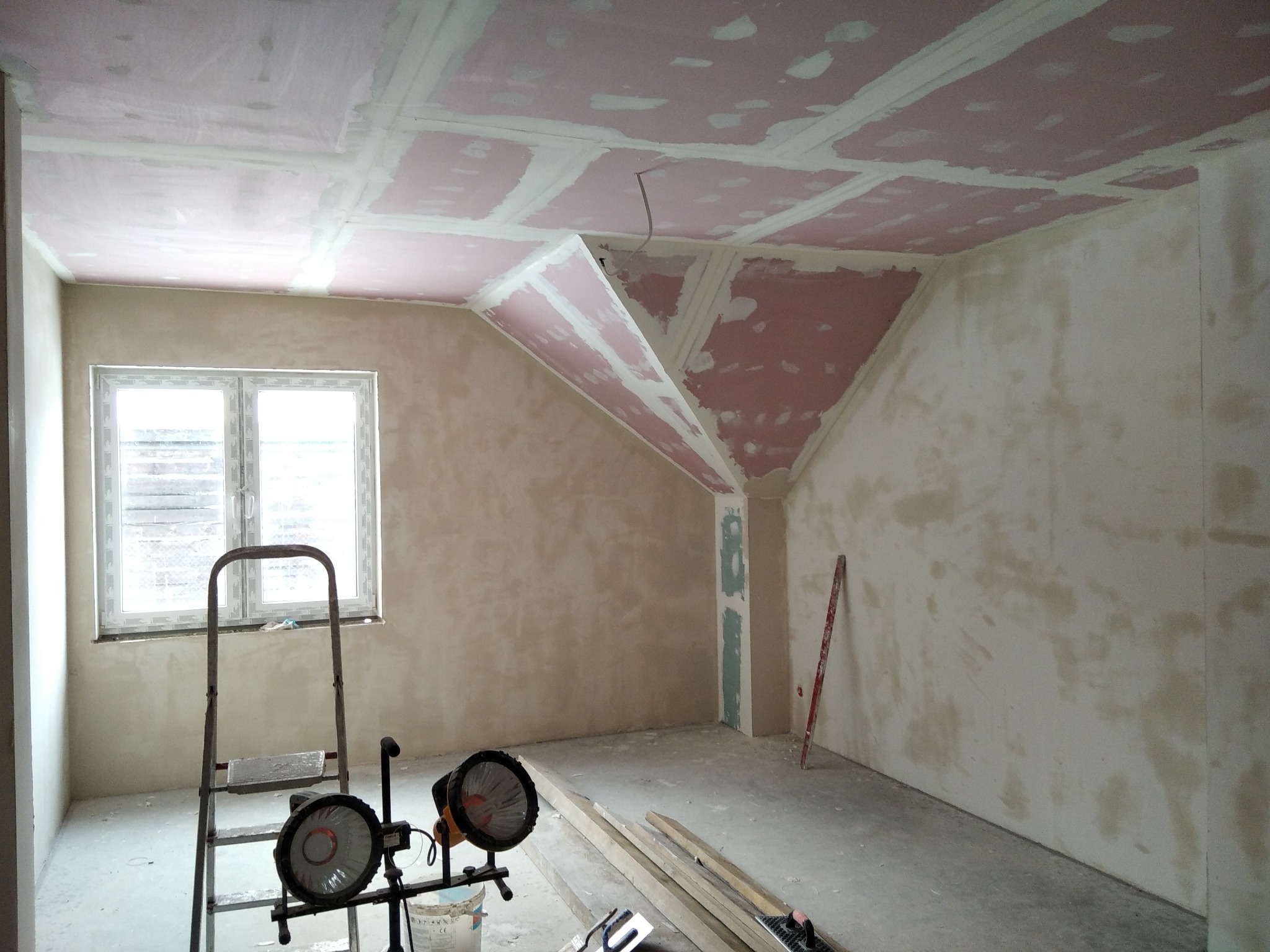 Pokój w trakcie prac wykończeniowych: ściany z nałożoną warstwą szpachli, sufit z płyt karton-gips z widocznymi łączeniami, drabina i lampy halogenowe w pomieszczeniu.