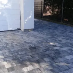 Nowo ułożona, szara kostka brukowa o prostokątnym kształcie, prowadząca do białej bramy garażowej z widocznym logo Hormann, z fragmentem ogrodzenia w tle.