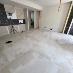 Vadzim Klysheika usługi remontowo-wykończeniowe - Wnętrze w trakcie prac wykończeniowych: ułożona marmurowa podłoga, ściany częściowo przygotowane pod gładzie, widoczny sprzęt oświetleniowy na statywie, listwy przypodłogowe w trakcie montażu.
