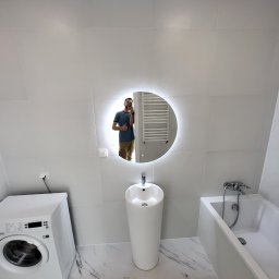 Vadzim Klysheika usługi remontowo-wykończeniowe - Nowoczesna łazienka z białymi płytkami, okrągłym lustrem z podświetleniem LED, wolnostojącą umywalką, wanną i pralką; odbicie mężczyzny robiącego zdjęcie w lustrze.