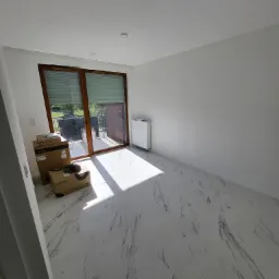 Pusty, nowo wykończony pokój z białymi ścianami, marmurową podłogą, grzejnikiem, oknem balkonowym z roletami i kartonami ze sprzętem AGD.