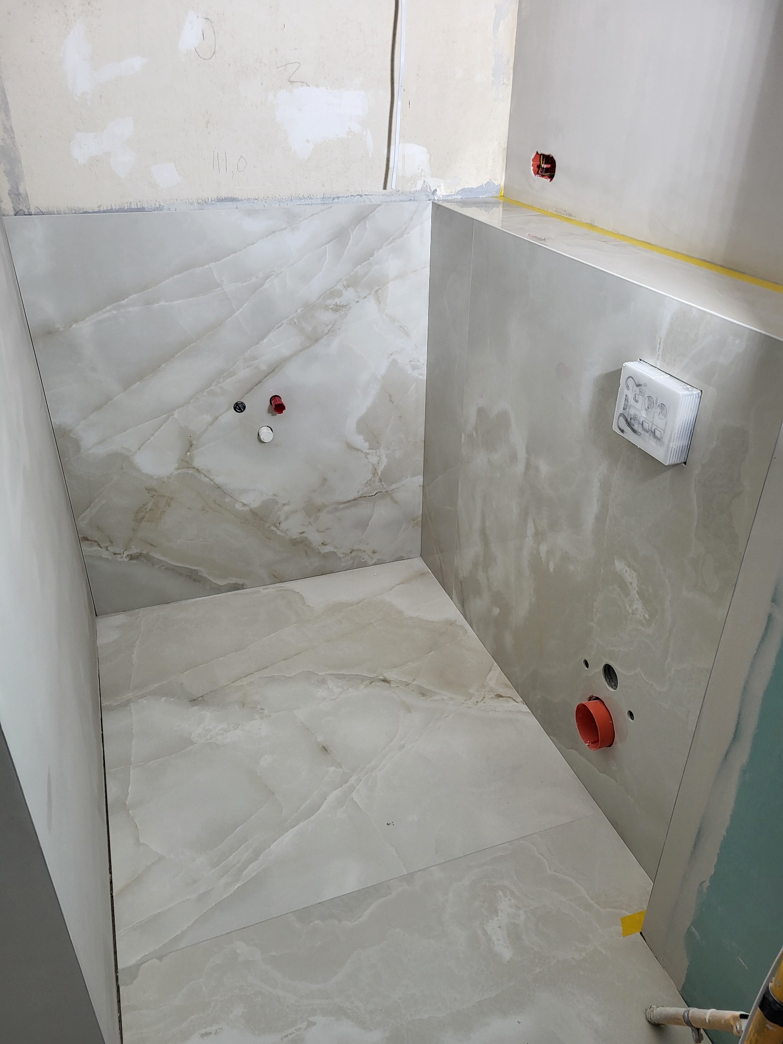 Wnętrze łazienki w trakcie prac wykończeniowych: ułożone płytki imitujące marmur na ścianach i podłodze, widoczne instalacje wodno-kanalizacyjne oraz fragmenty surowych ścian przygotowywanych...