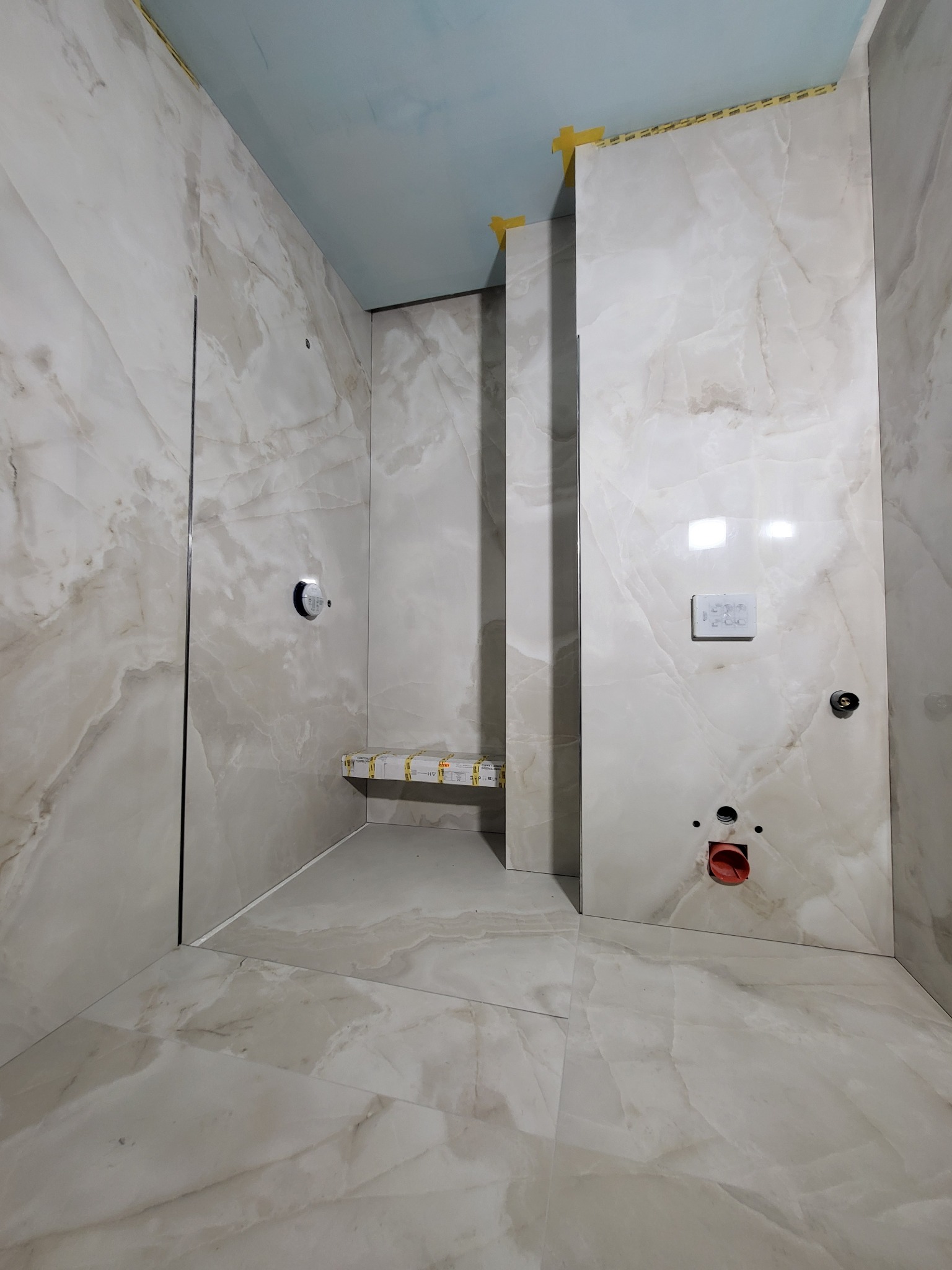 Łazienka w trakcie wykańczania: marmurowe płytki na ścianach i podłodze, widoczne otwory na armaturę, fragment siedziska zabezpieczony folią, taśma maskująca na górze płytek.