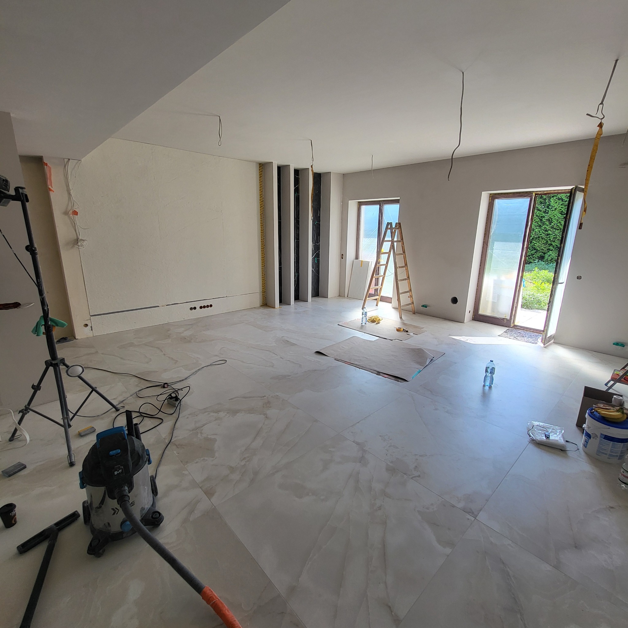 Przestronne wnętrze w trakcie prac wykończeniowych: jasna podłoga z płytek imitujących marmur, ściany przygotowane pod malowanie, widoczne narzędzia, drabina oparta o ścianę, okna zabezpieczone folią.
