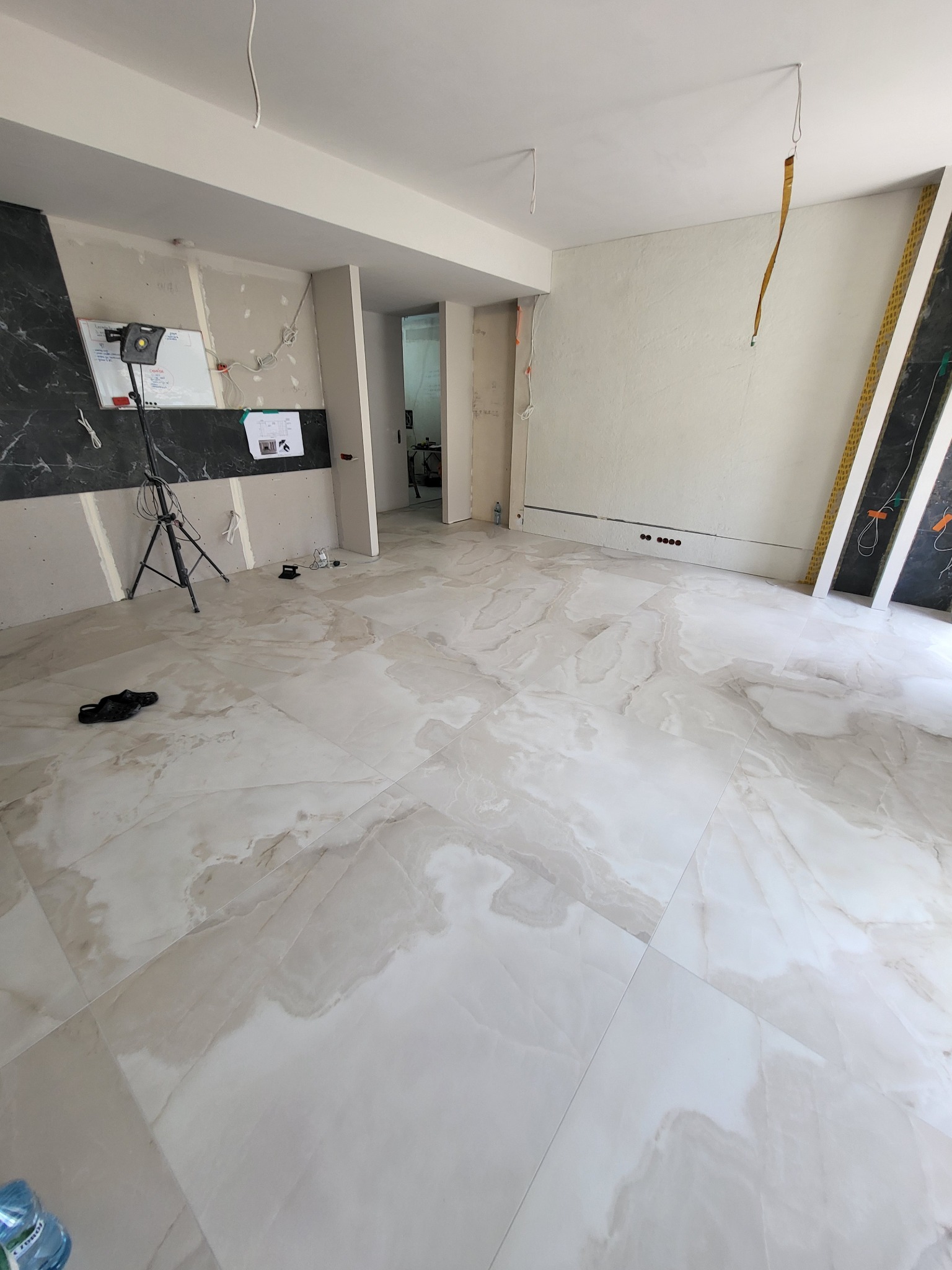 Wnętrze w trakcie prac wykończeniowych: ułożona marmurowa podłoga, ściany częściowo przygotowane pod gładzie, widoczny sprzęt oświetleniowy na statywie, listwy przypodłogowe w trakcie montażu.