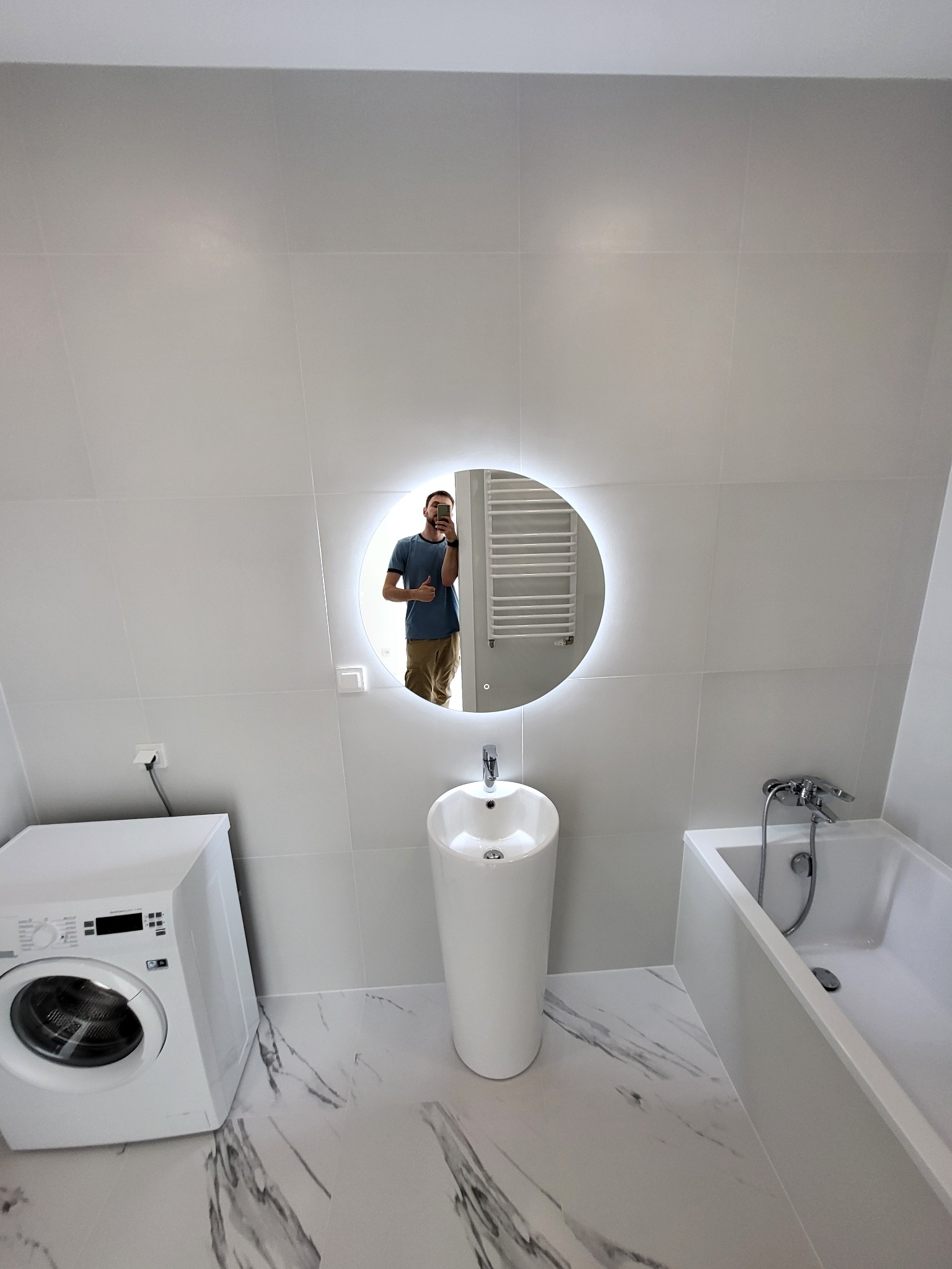 Nowoczesna łazienka z białymi płytkami, okrągłym lustrem z podświetleniem LED, wolnostojącą umywalką, wanną i pralką; odbicie mężczyzny robiącego zdjęcie w lustrze.