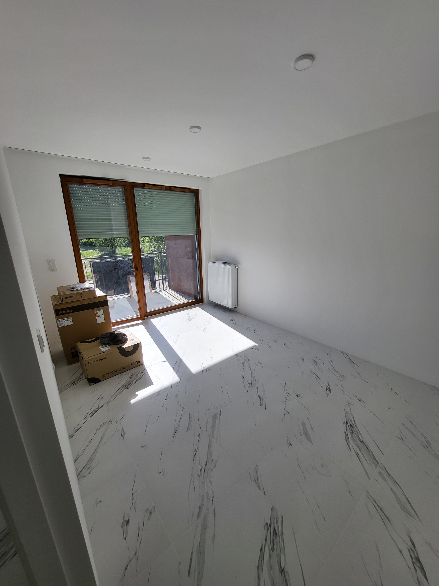 Pusty, nowo wykończony pokój z białymi ścianami, marmurową podłogą, grzejnikiem, oknem balkonowym z roletami i kartonami ze sprzętem AGD.