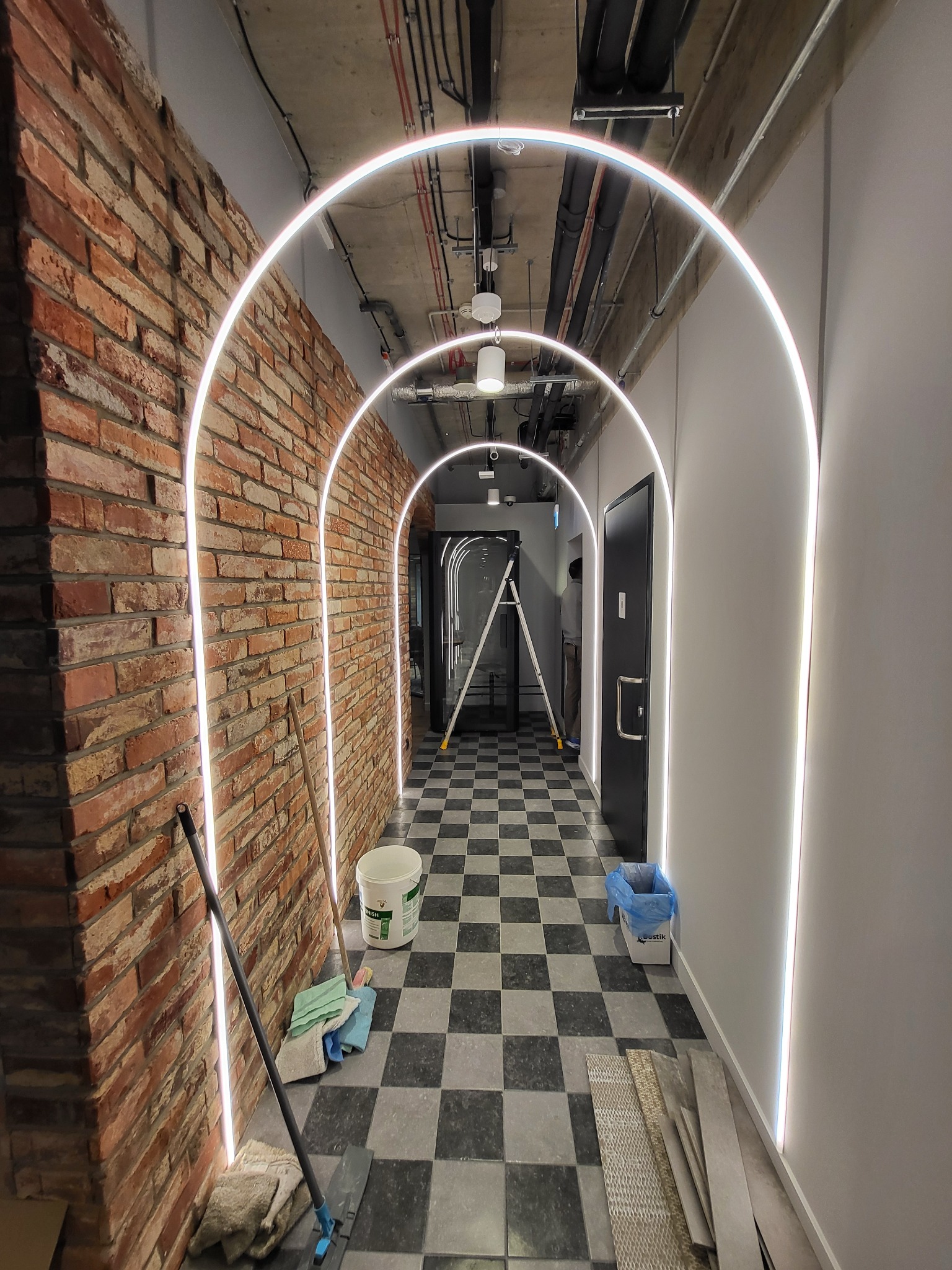 Korytarz z wykończonymi ścianami z cegły i białych paneli, oświetlony łukami LED, podłoga w szachownicę, widoczne narzędzia malarskie i drabina w tle.