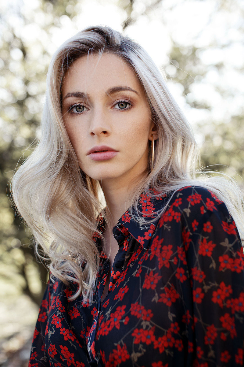 Portret kobiety z blond włosami w kwiecistej sukience, z delikatnym makijażem i złotymi kolczykami na tle rozmytej zieleni.