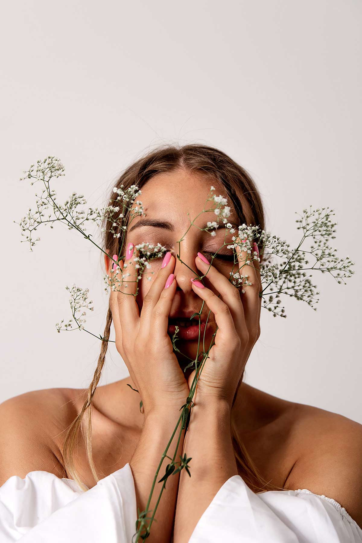 Portret kobiety z zamkniętymi oczami, ozdobionej gałązkami gipsówki, dłonie z różowym manicure zakrywają częściowo twarz, w tle białe tło.