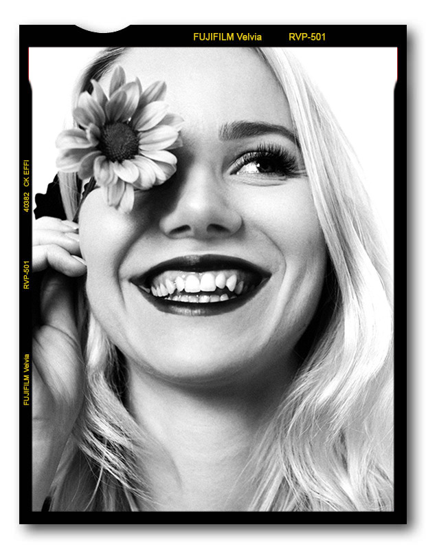 Portret uśmiechniętej blondynki z kwiatem we włosach, w czarno-białej stylistyce, imitujący klatkę filmu Fujifilm Velvia.