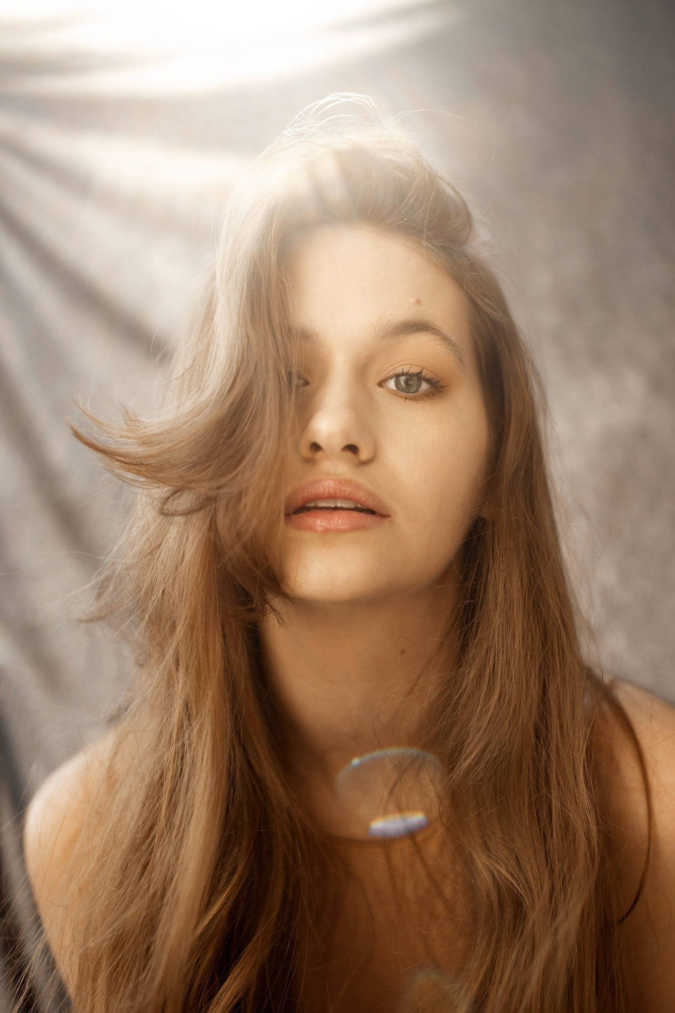Portret młodej kobiety z długimi, brązowymi włosami, częściowo zakrywającymi twarz, z widocznym blaskiem słonecznym w tle i odblaskami na włosach.
