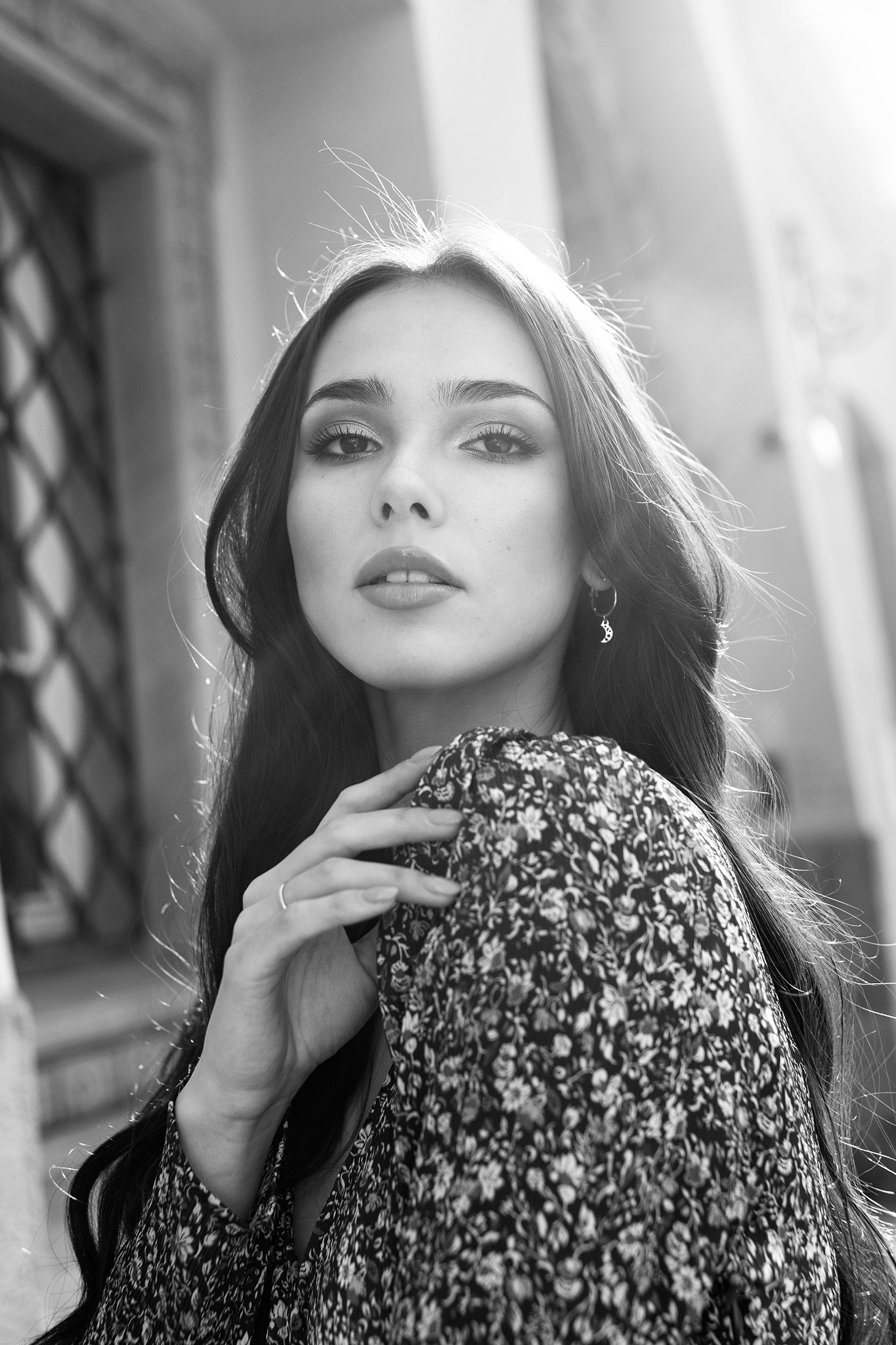 Portret młodej kobiety z długimi, ciemnymi włosami, w kwiecistej sukience, z delikatnym makijażem i srebrnym kolczykiem w kształcie księżyca, sfotografowanej w czerni i bieli z niskiego kąta.