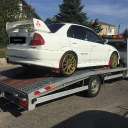 Biały sportowy samochód Mitsubishi Lancer Evolution VI z dużym spojlerem, złotymi felgami i naklejkami Ralliart umieszczony na platformie lawety samochodowej z włączonymi pomarańczowymi światłami...